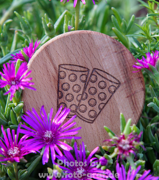 Holzdeckel für Dubbeglas mit eingeprägten Gläsern, umgeben von blühenden Pflanzen.