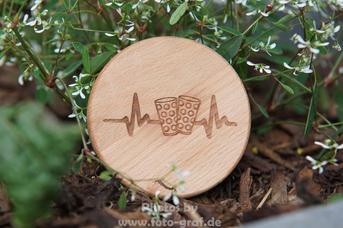 Stylischer Dubbedeckel aus Buchenholz mit Glas- und Herzschlag-Symbol auf einer Blumenwiese.
