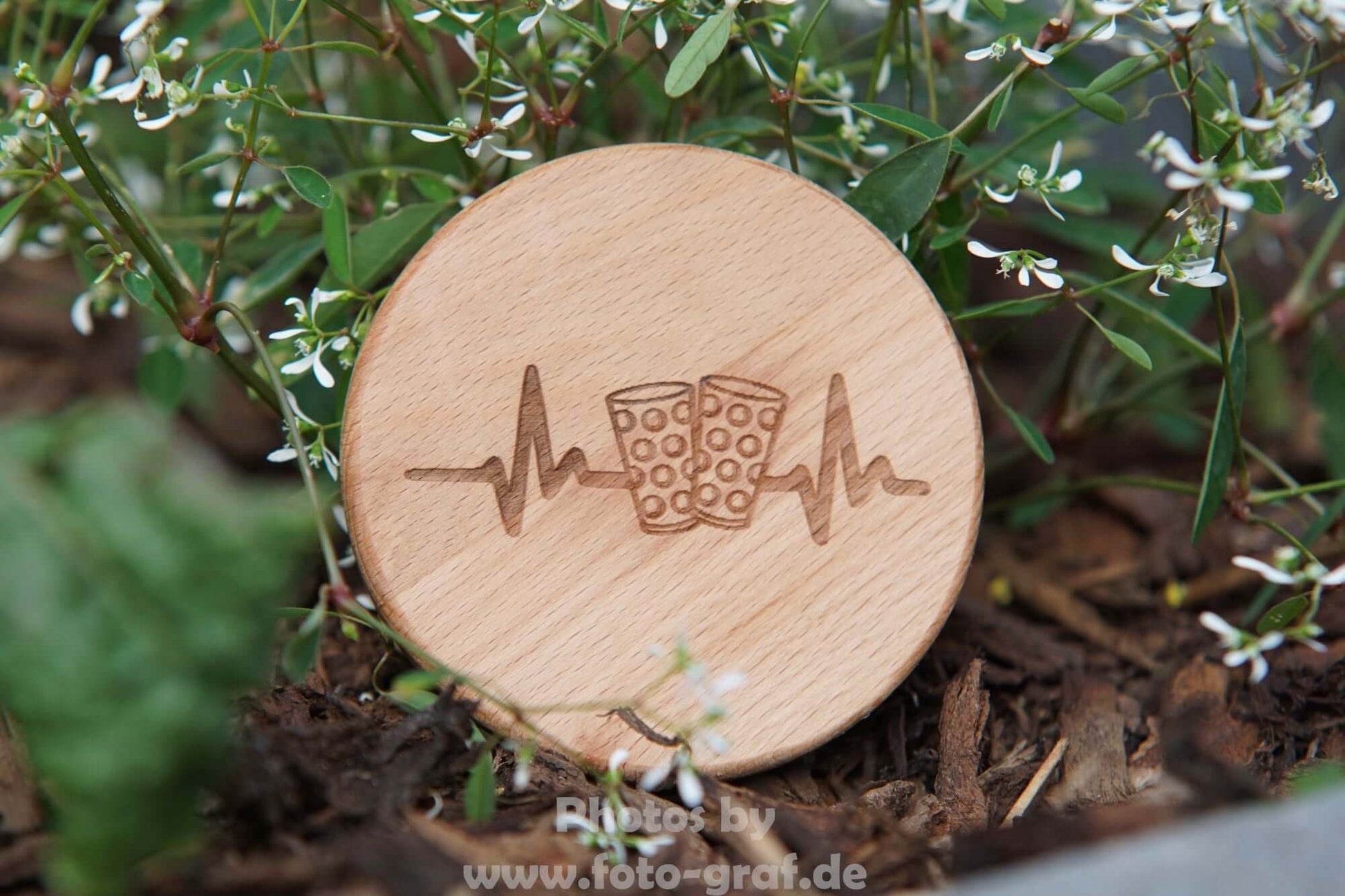 Stylischer Dubbedeckel aus Buchenholz mit Glas- und Herzschlag-Symbol auf einer Blumenwiese.